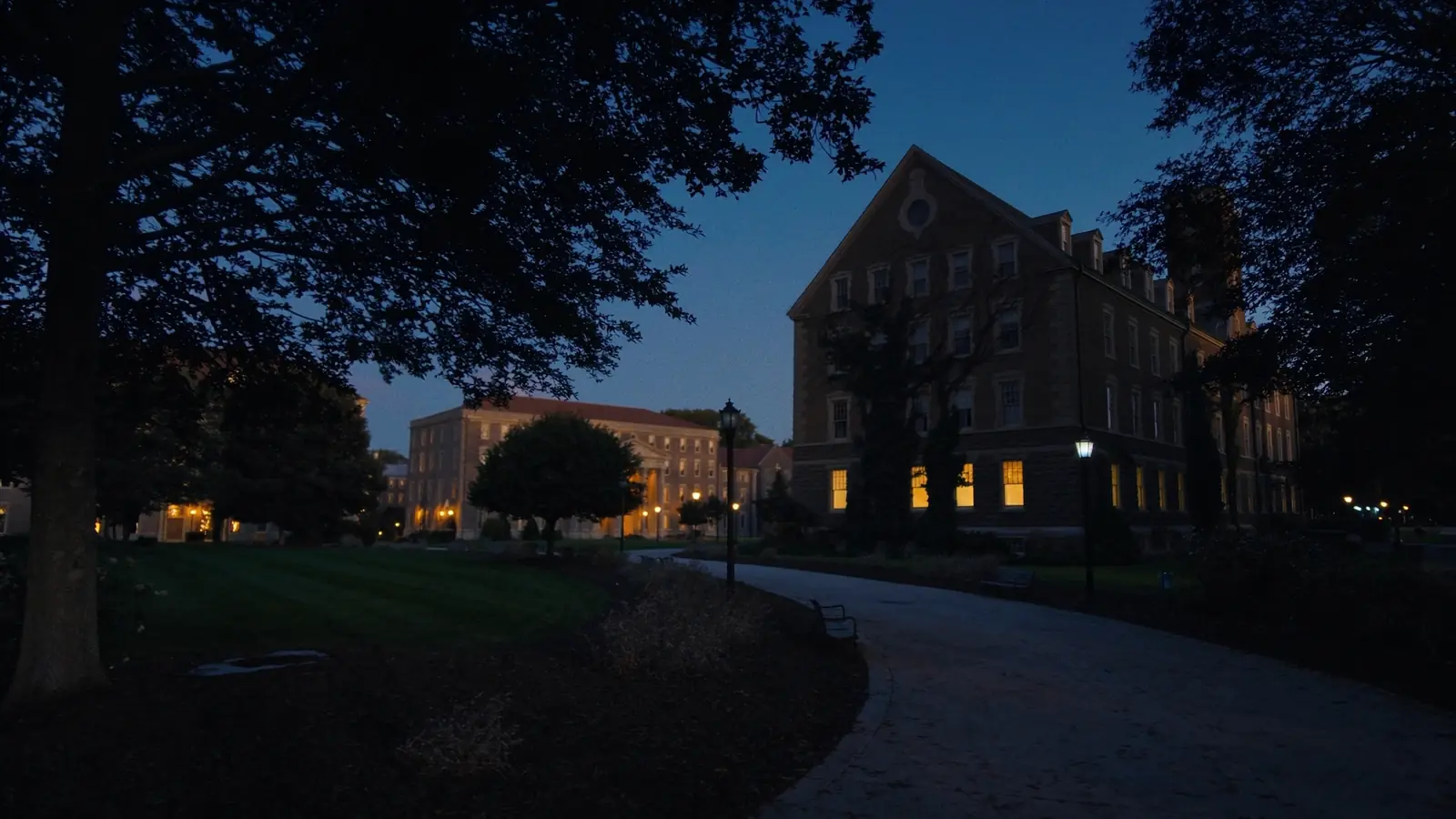 A quiet university campus walkway at dusk with lit buildings and empty benches